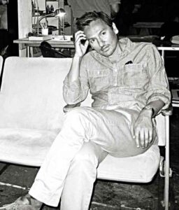 Black and white portrait of Ramon Garcia sitting on white sofa, holding head with a cocked arm, elbow resting on knee. 