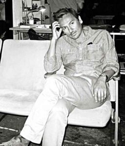 Black and white portrait of Ramon Garcia sitting on white sofa, holding head with a cocked arm, elbow resting on knee.