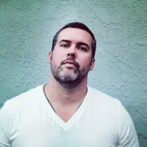 Poet Luke Johnson in a white v-neck shirt, standing against a light green/grey background