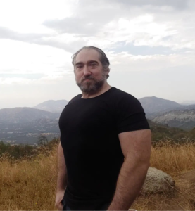 Headshot of Michael Meyerhofer in a black short-sleeved t-shirt, standing in front of foggy-topped mountains in the distance with golden grass in the immediate background. 