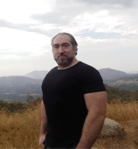 Headshot of Michael Meyerhofer in a black short-sleeved t-shirt, standing in front of foggy-topped mountains in the distance with golden grass in the immediate background.