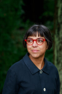 Color portrait of poet Judy Halebsky, looking to the left in red-frame glasses and a navy blue top with green blurred background