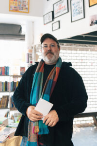 Portrait of Caleb Nichols with a beard, wearing a hat, jacket, and scarf inside an interior space. 